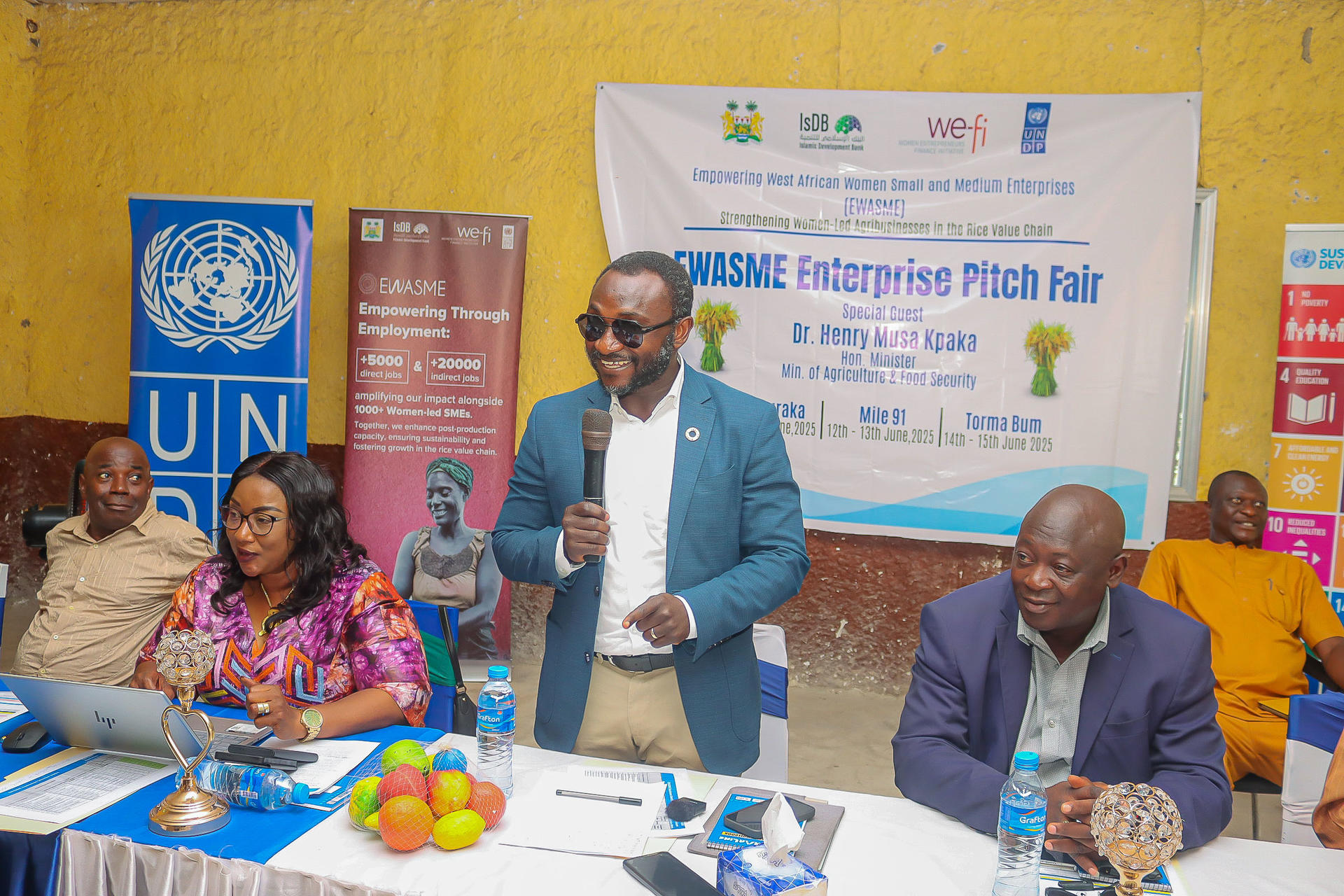A speaker presents at a pitch event, with attendees at tables and banners in the background.