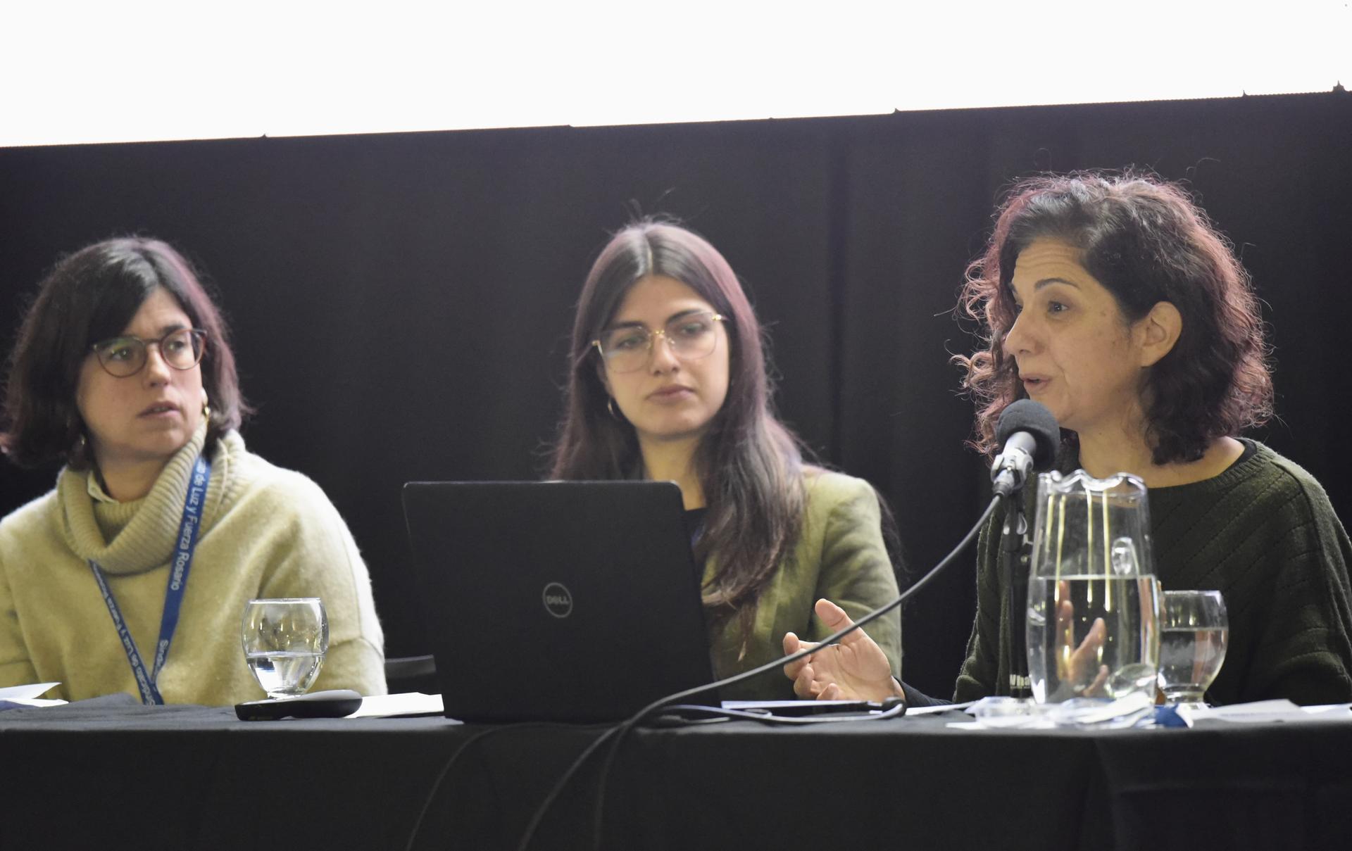 Tres mujeres sentadas en un panel de discusión, con una computadora y vasos de agua en la mesa.