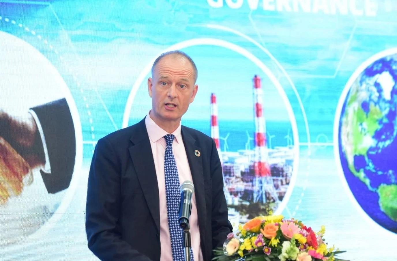 A man in a suit speaks at a podium with flowers, colorful graphics in the background.