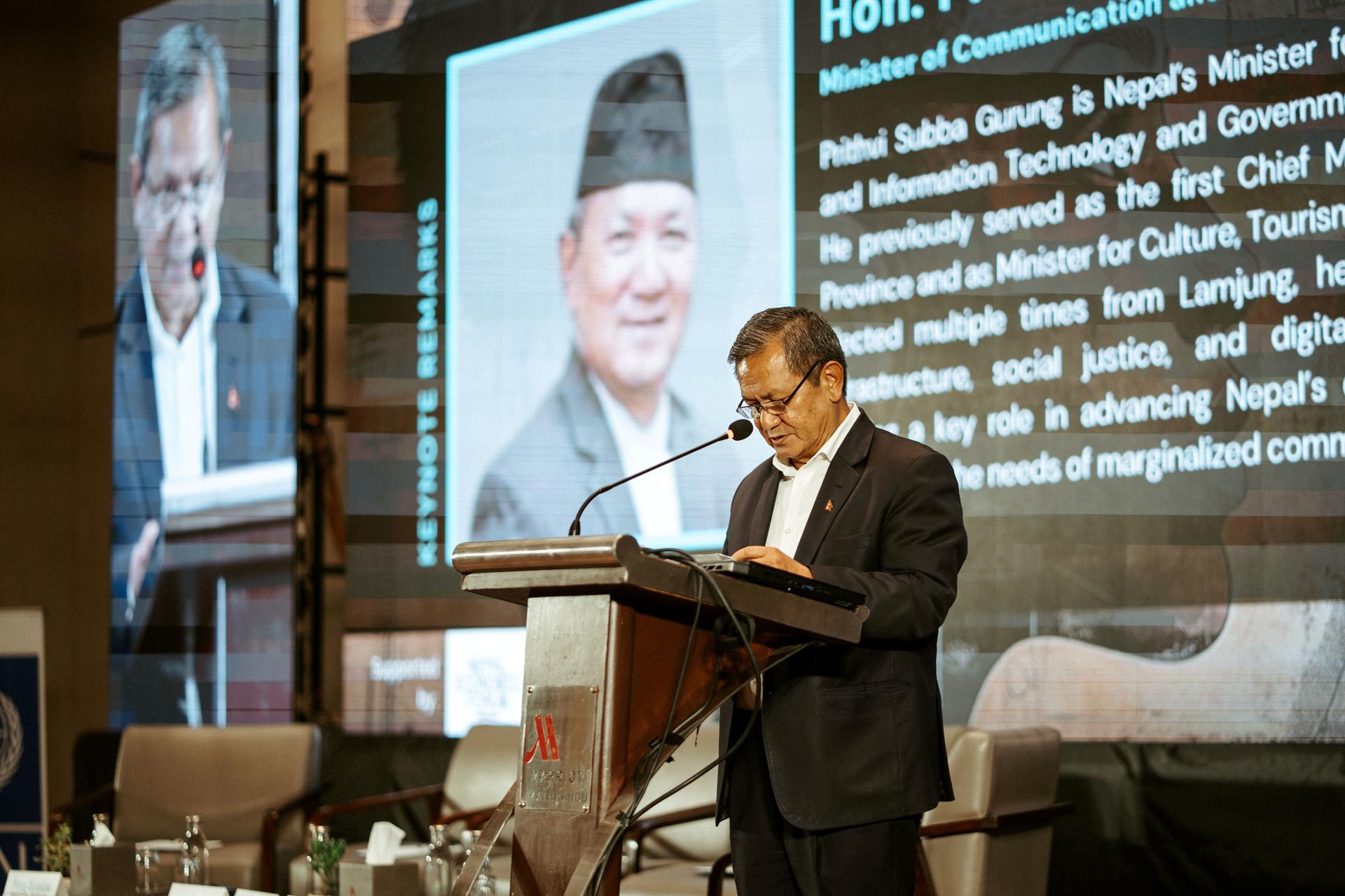 A man stands at a podium speaking, with a large screen displaying another man's image behind him.