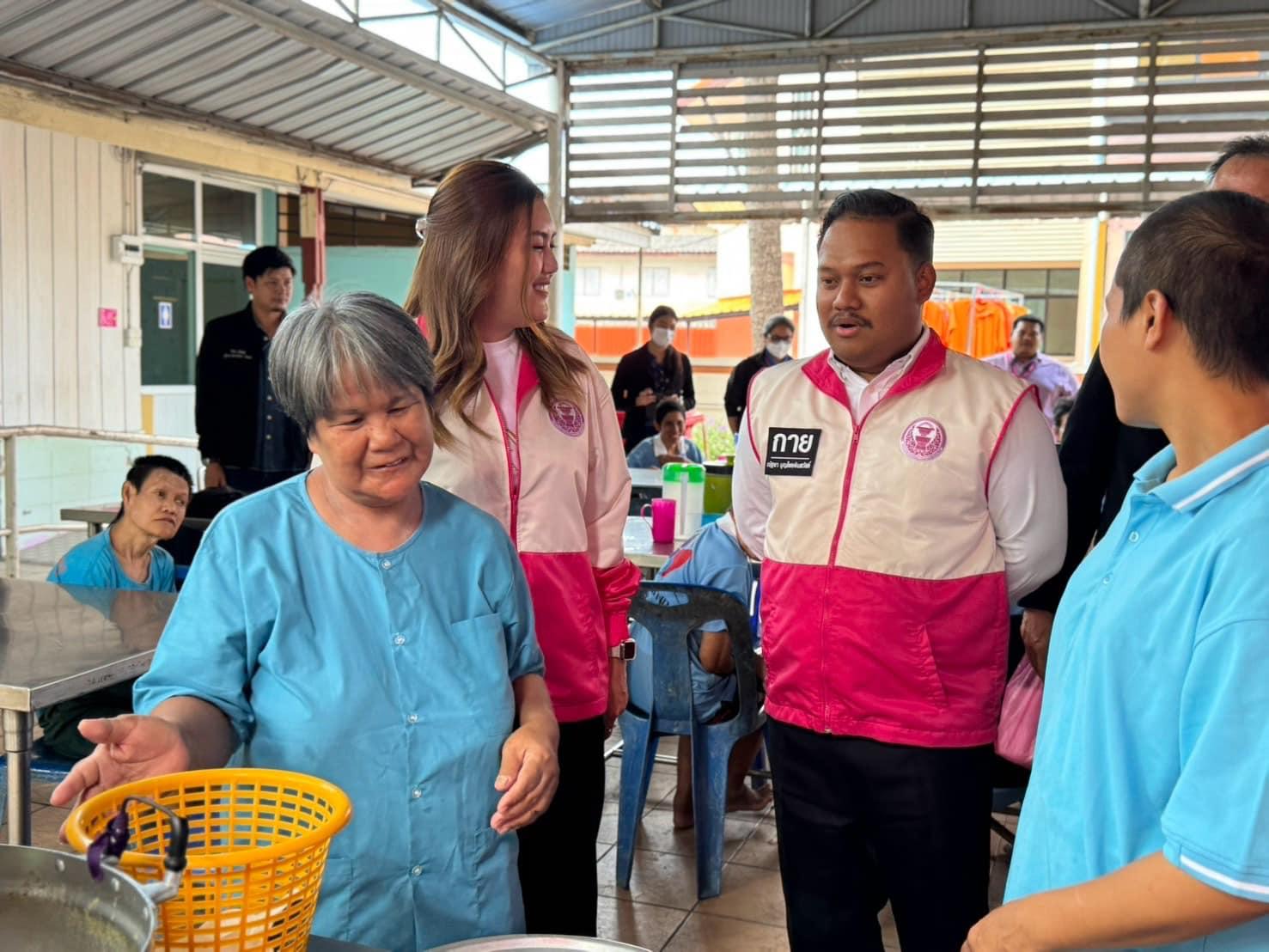 A smiling woman in blue interacts with two individuals in pink vests at a communal kitchen.