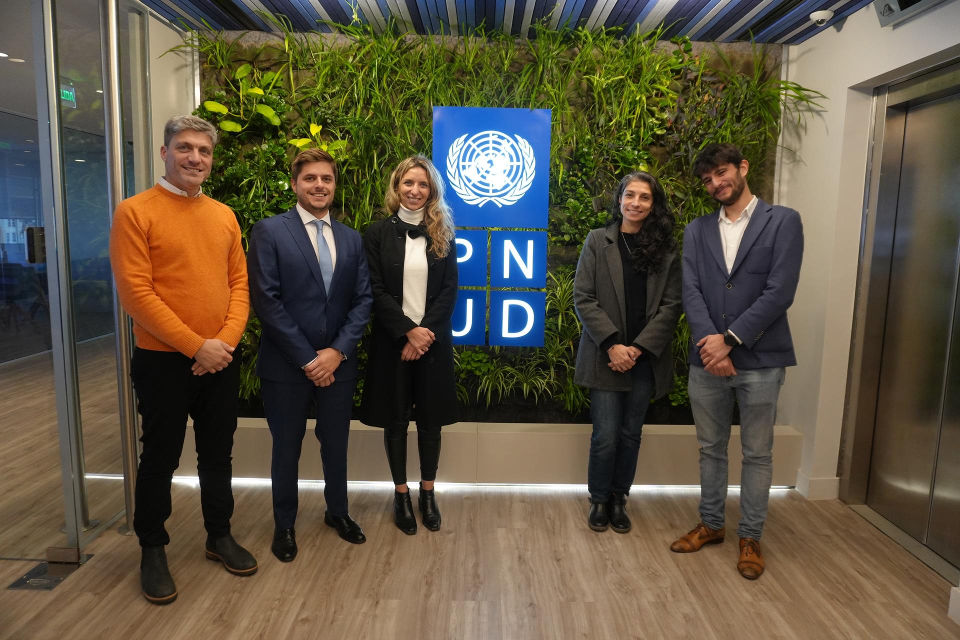 Cinco profesionales parados frente a una pared con plantas verdes con el logo de PNUD en el centro.