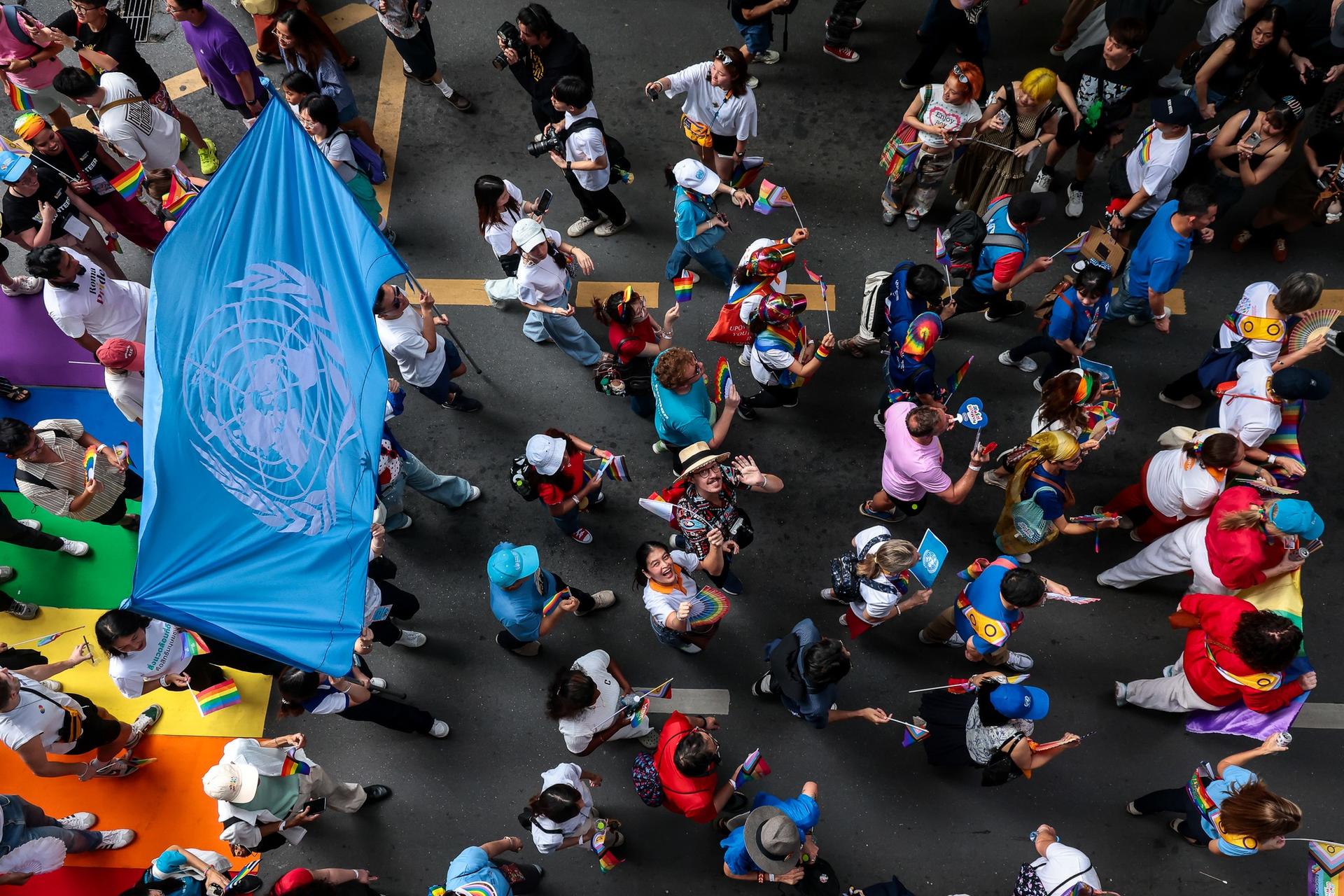 Aerial view of a diverse crowd at a parade, featuring a blue UN flag and colorful road decorations.
