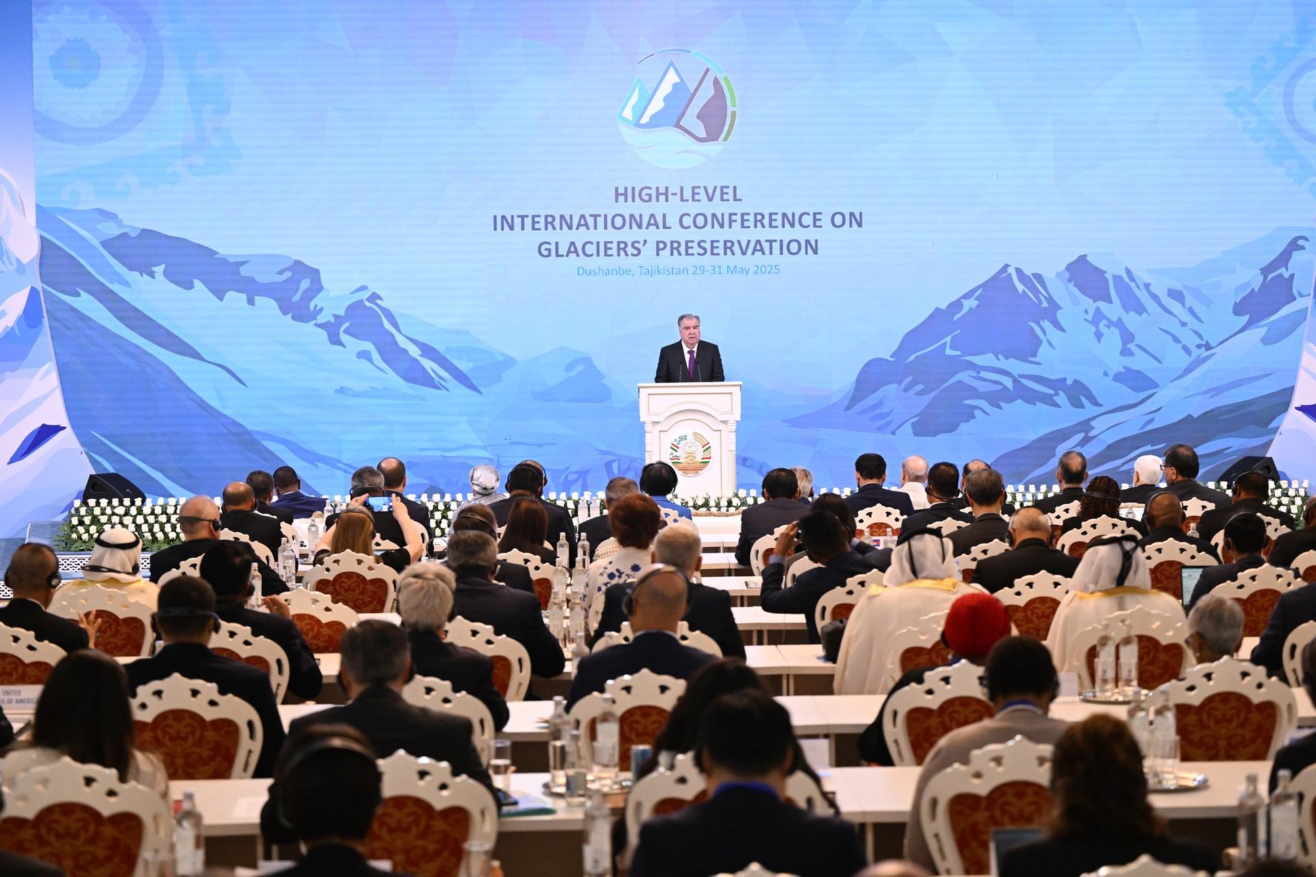 A speaker addresses an audience at an international conference, with scenic mountains in the background.