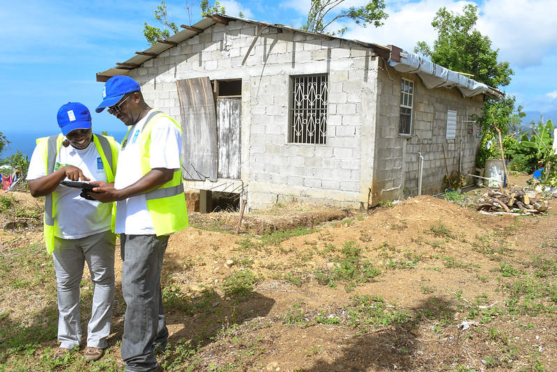 Two workers in safety vests and caps review a tablet near a small, weathered house.