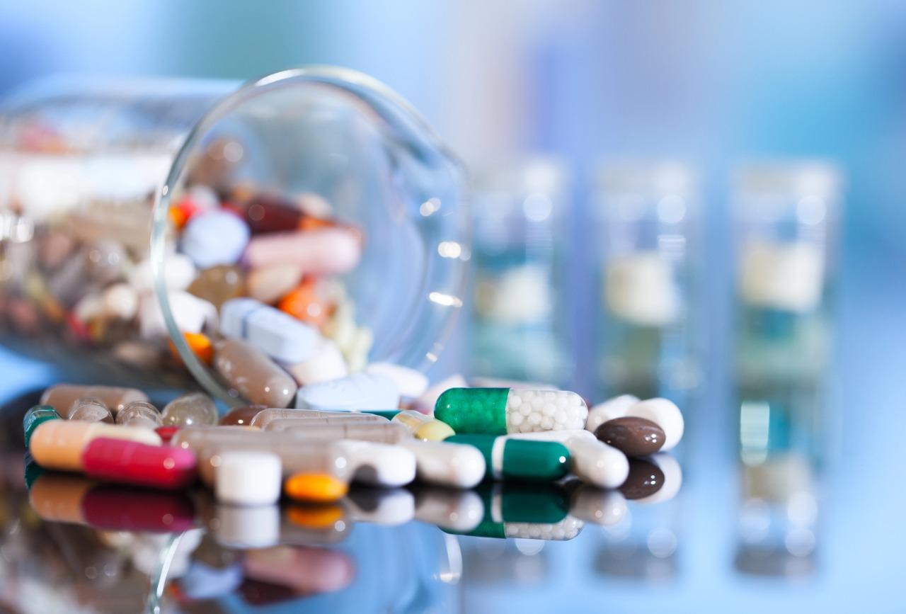 Scattered capsules and tablets next to a tipped-over glass jar, with blurred vials in the background.