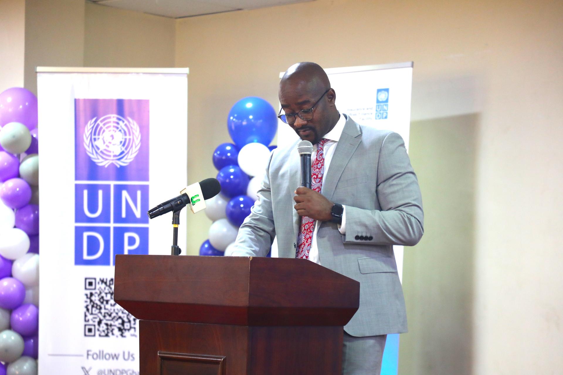 A man in a gray suit speaks at a podium with UNDP branding, surrounded by balloons.