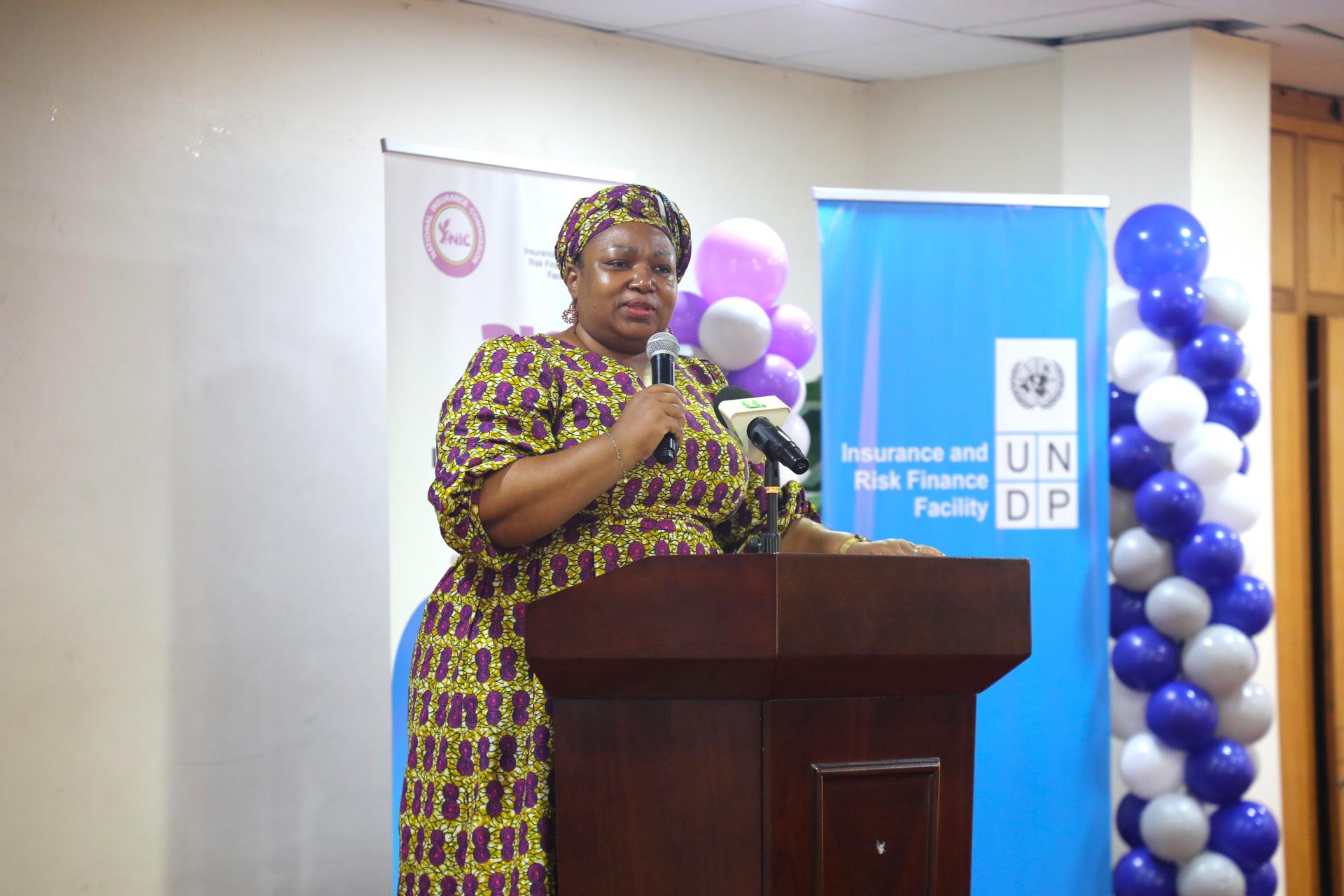 A woman in a patterned dress speaks at a podium, with banners and balloons in the background.