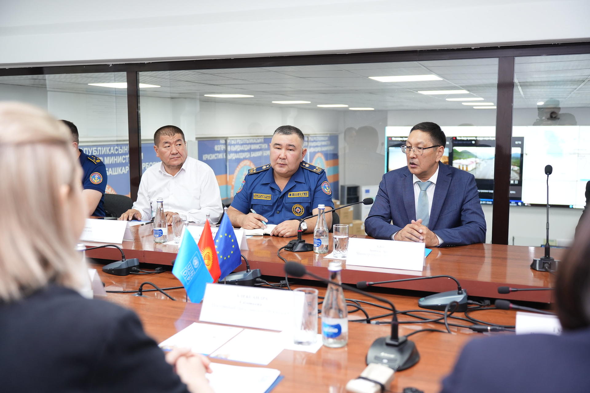 A conference room with officials discussing at a table, flags and documents present.