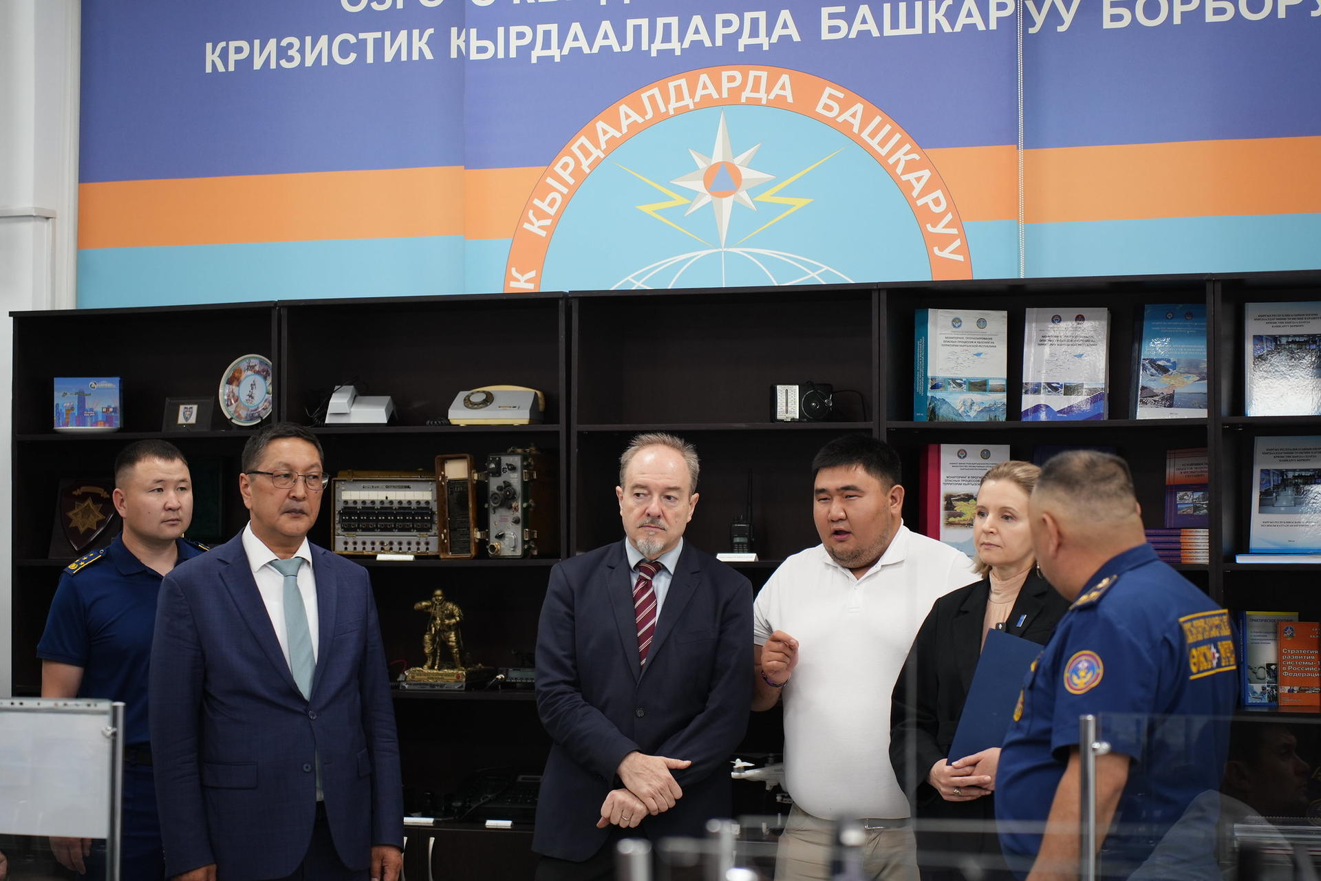 Group of officials interacting inside an office with awards and documents in the background.
