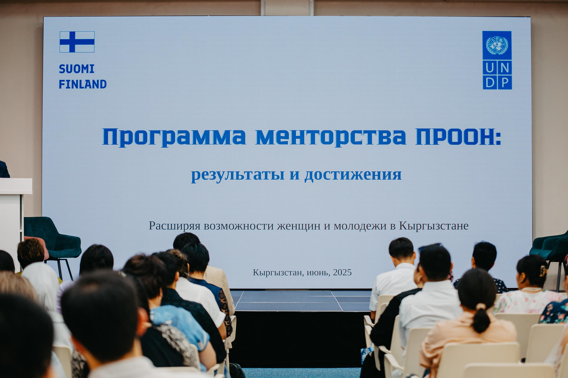 Audience members seated facing a stage with a large presentation screen displaying text in Russian and logos.