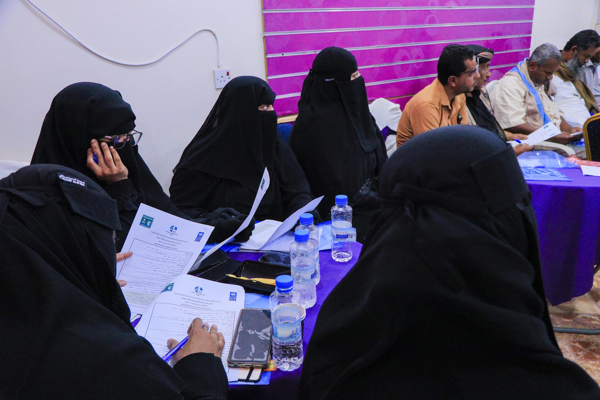 Women in hijabs seated at a table, engaged in discussion with others in a meeting.