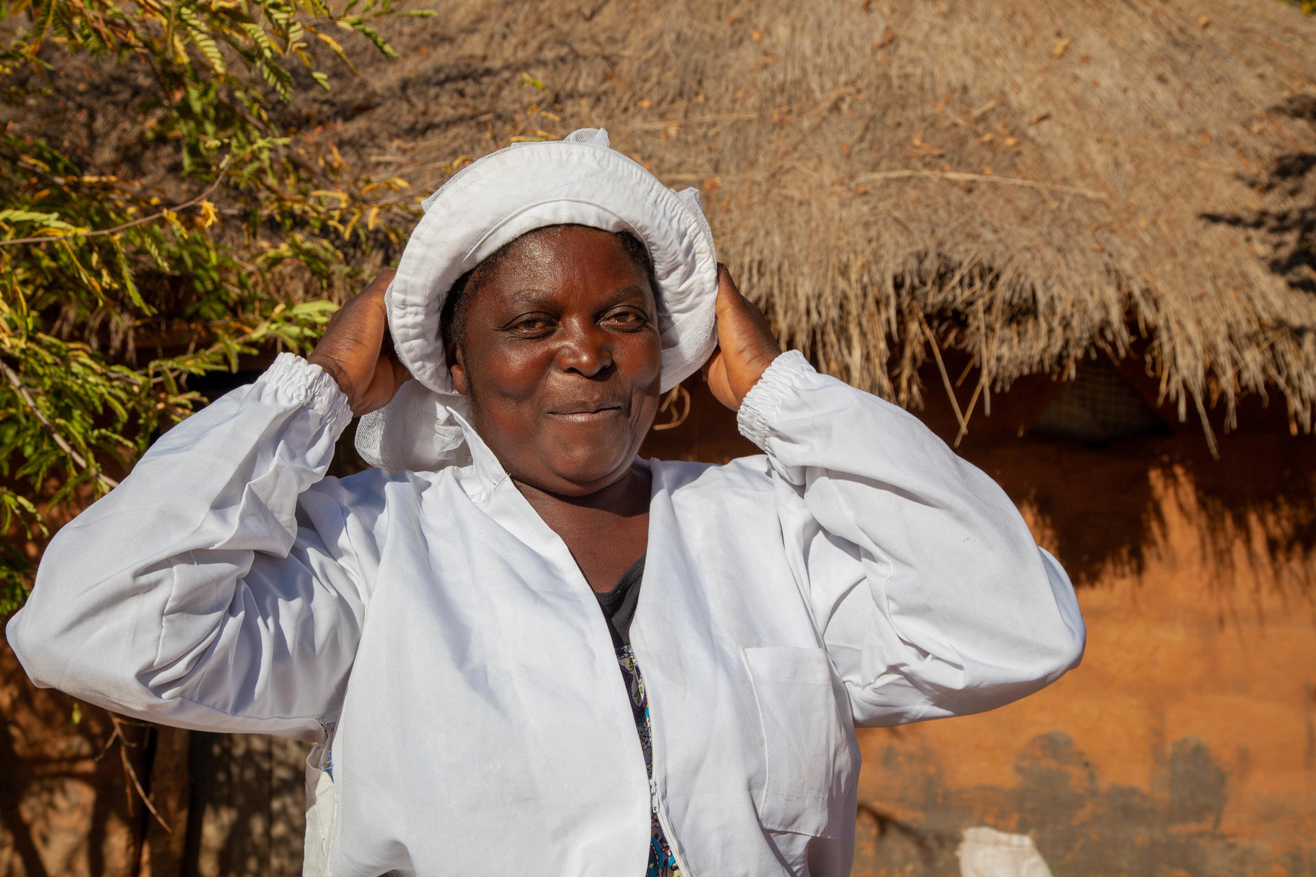 Smiling woman in a white coat adjusts a head wrap outdoors near a thatched roof.