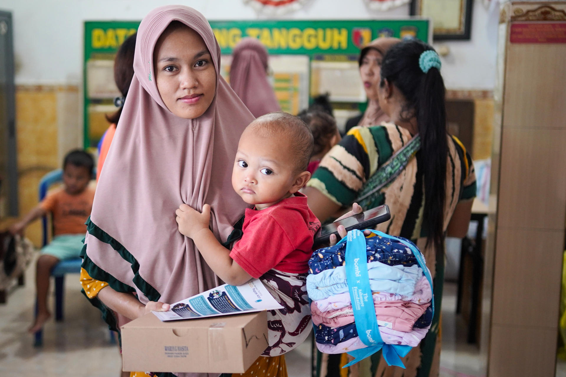 Woman wearing a hijab holds a child and a box in a busy indoor setting.
