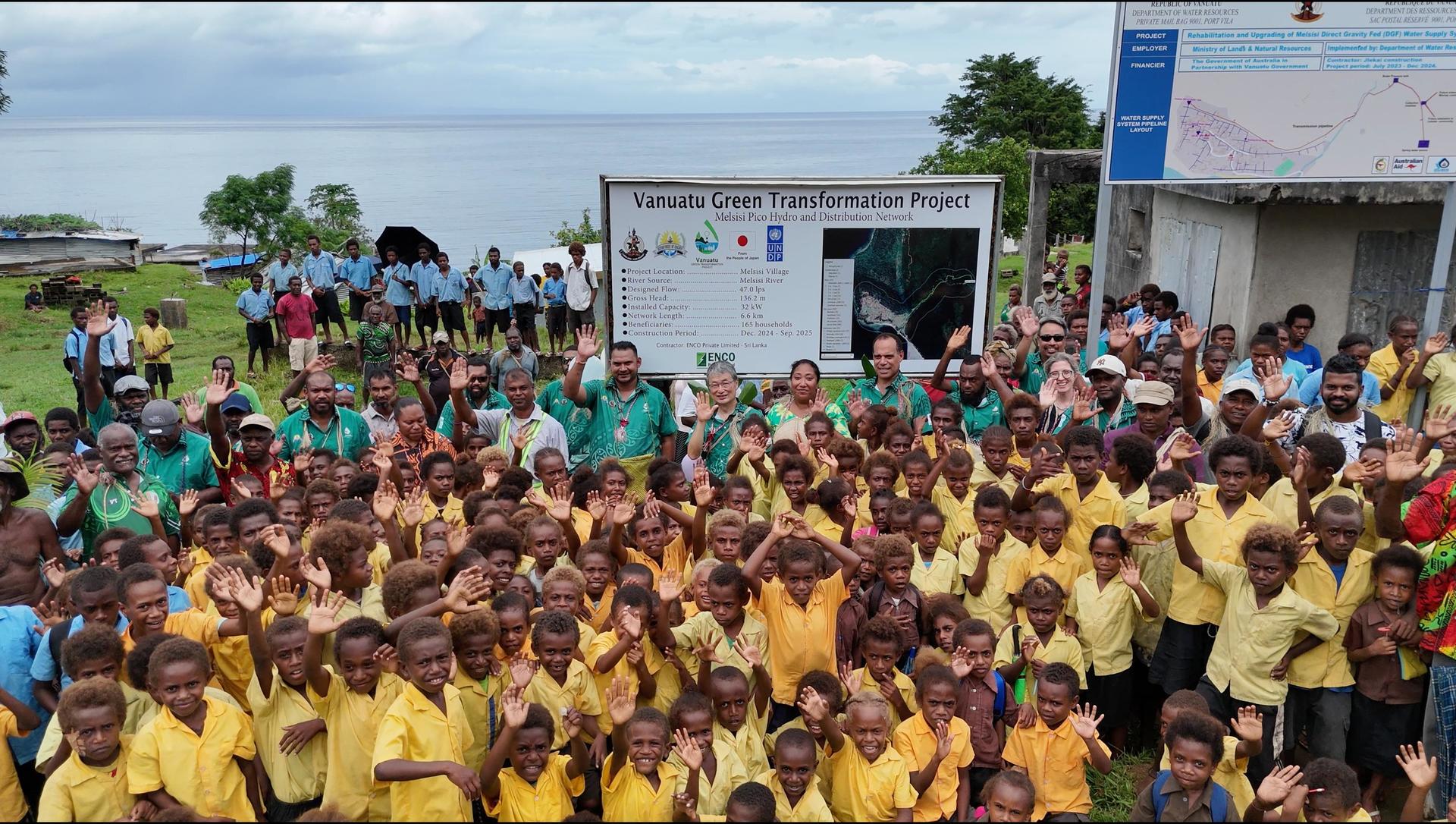 A large group of children in yellow shirts cheerfully wave their hands outdoors.