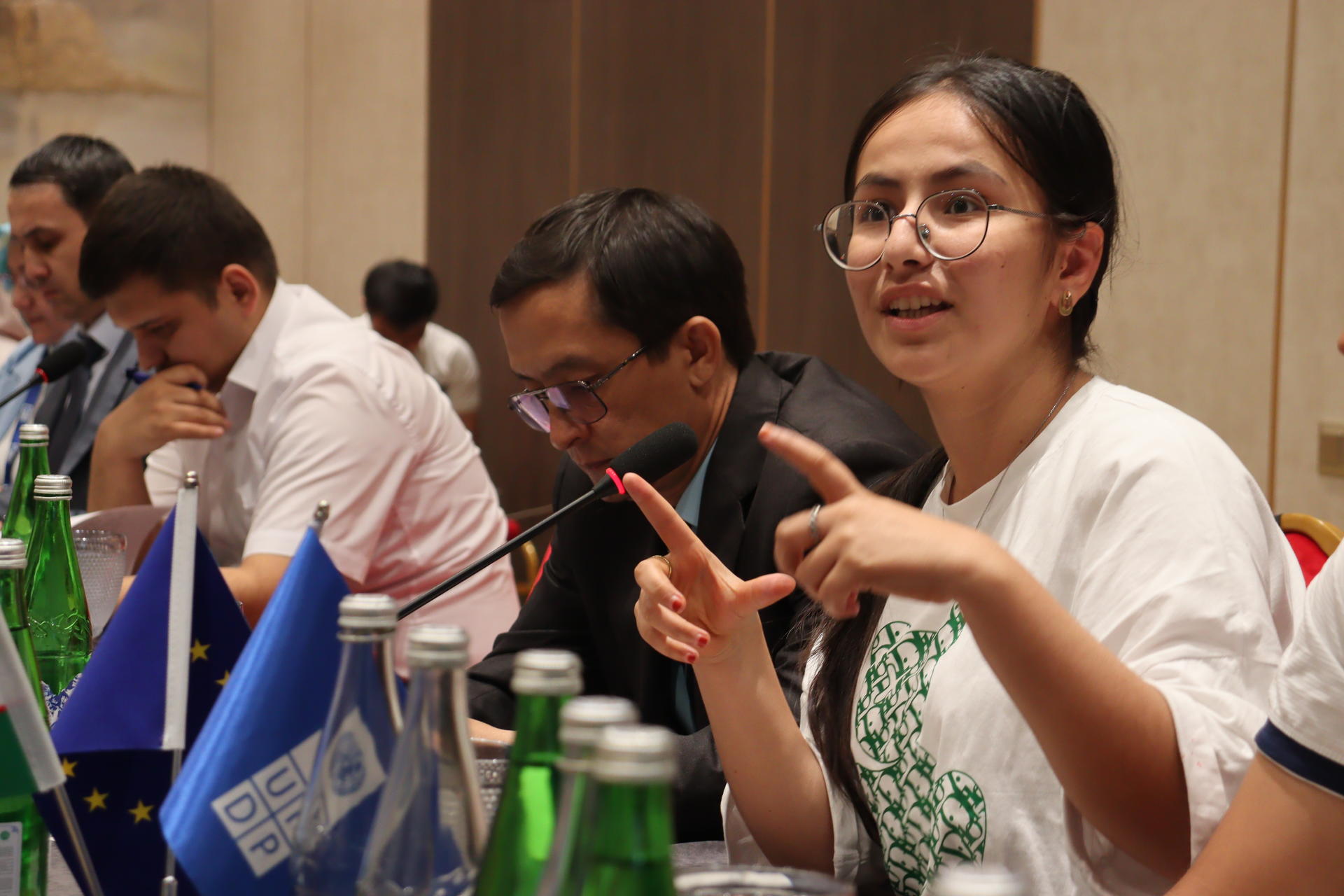A young woman gestures while speaking at a conference table, with participants and flags in view.