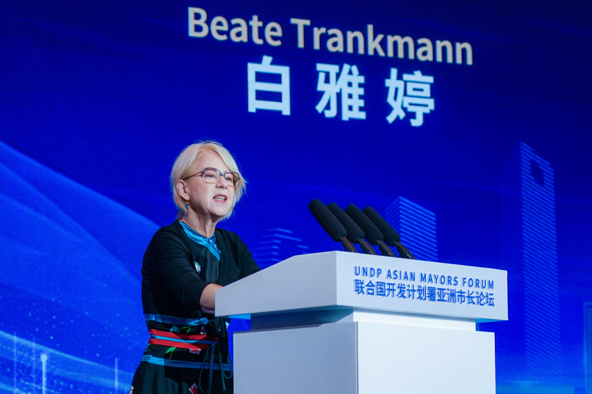 A woman speaks at a podium with a blue backdrop featuring her name in English and Chinese.