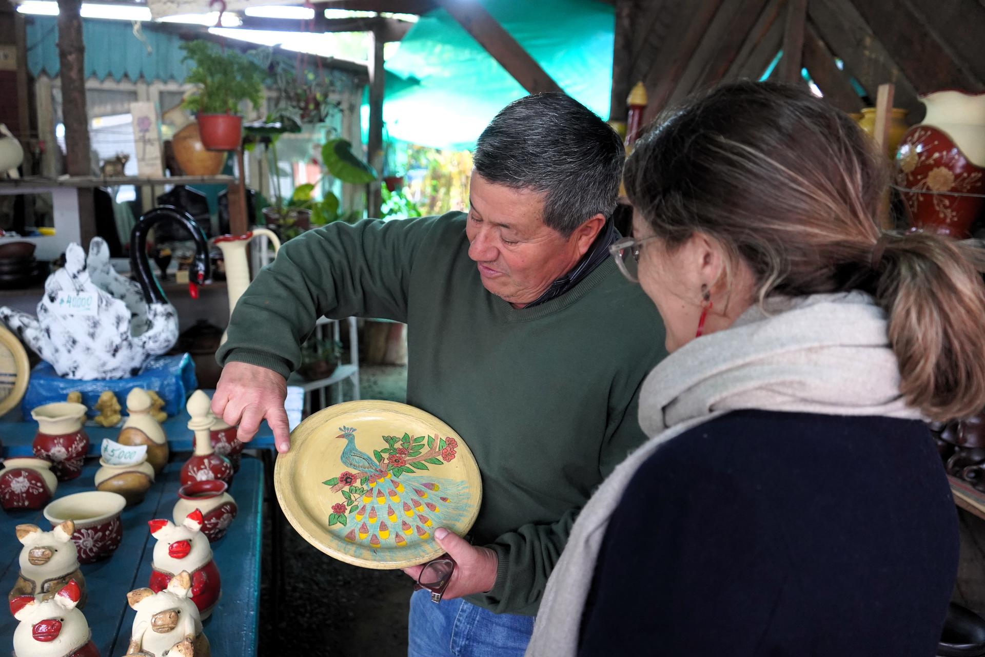 A vendor shows a colorful, decorated plate to a woman at a market filled with pottery.