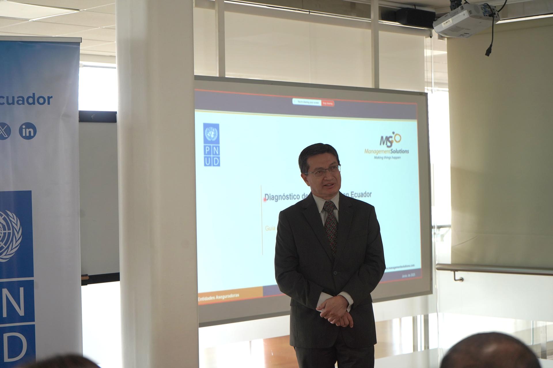 A man in a suit stands in front of a presentation screen in a modern office setting.