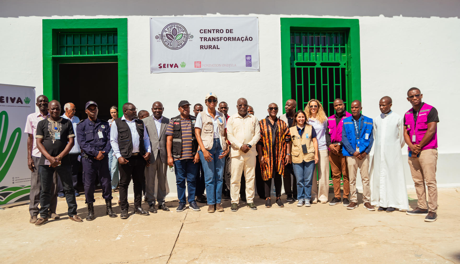 Group photo of diverse individuals in front of a building with green windows and a sign.