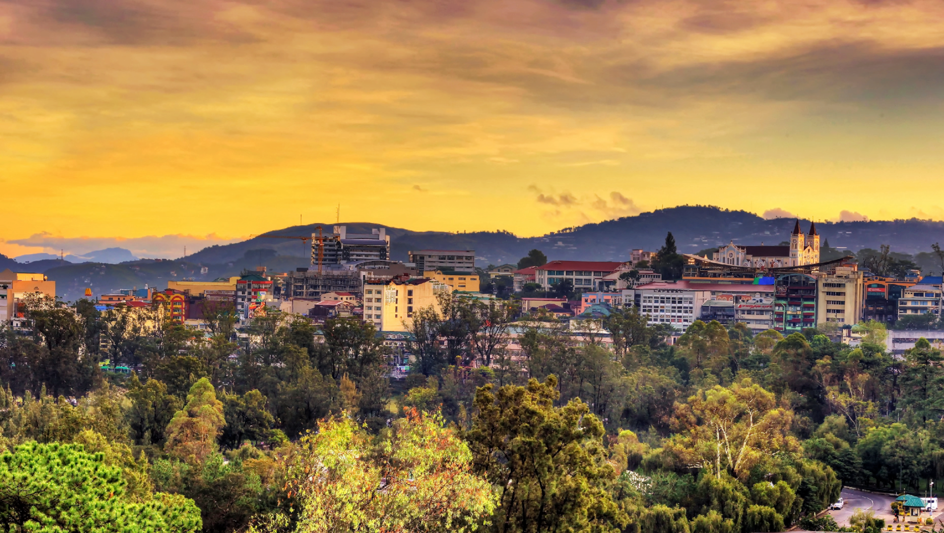 Panoramic view of a city at sunset, surrounded by hills and lush greenery.