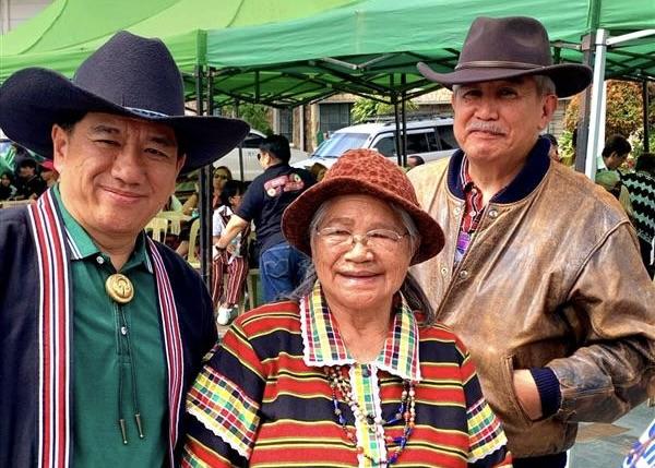 Three people smile in traditional attire, set against green market tents.