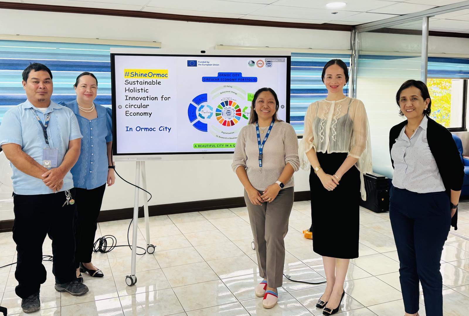 Five professionals pose in an office setting, smiling in front of a screen displaying graphics.