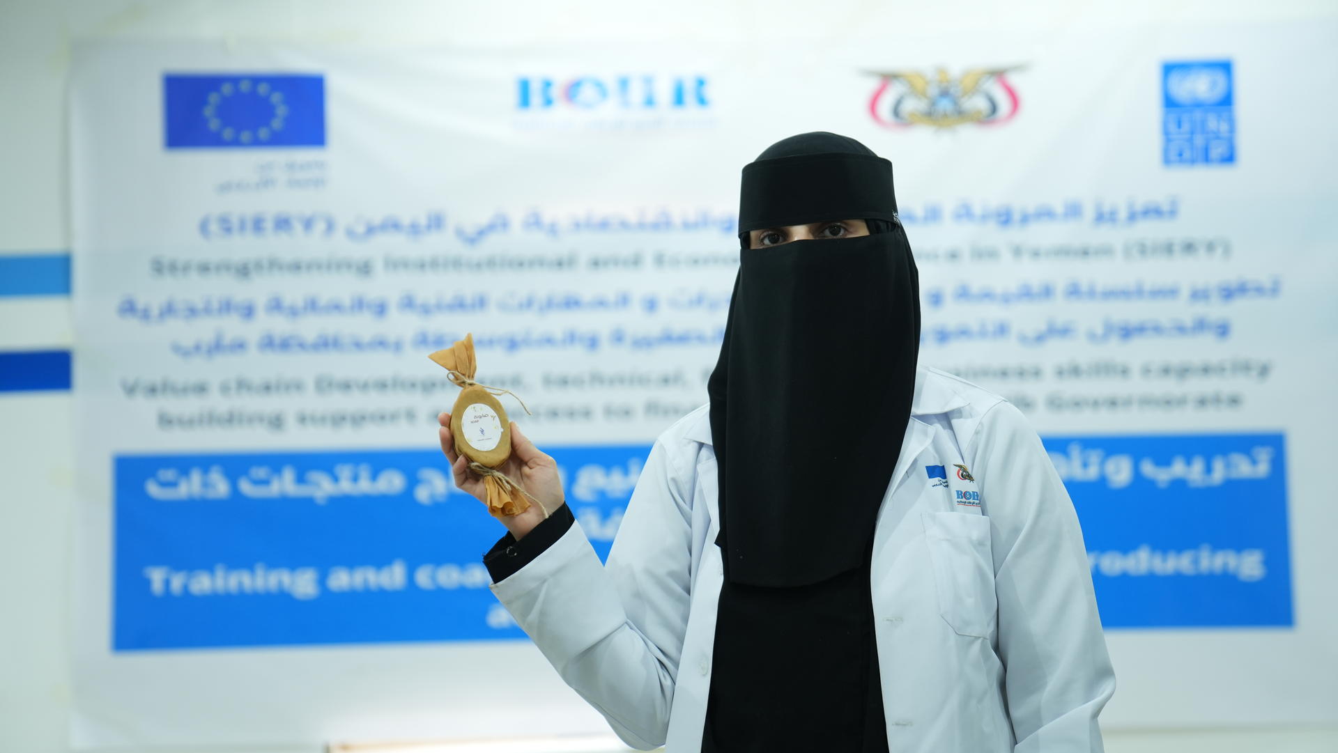 A woman wearing a niqab and lab coat holds an award in front of a banner.