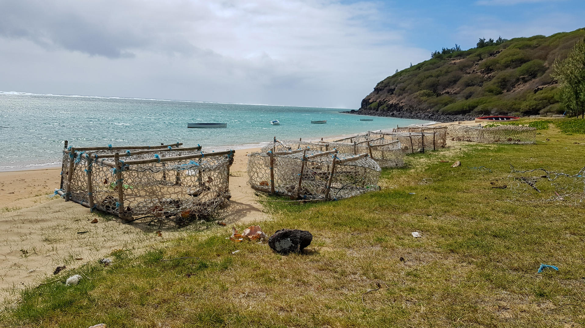 Coastal scene with cages on the beach, grass in the foreground, and a rocky hill in the background.