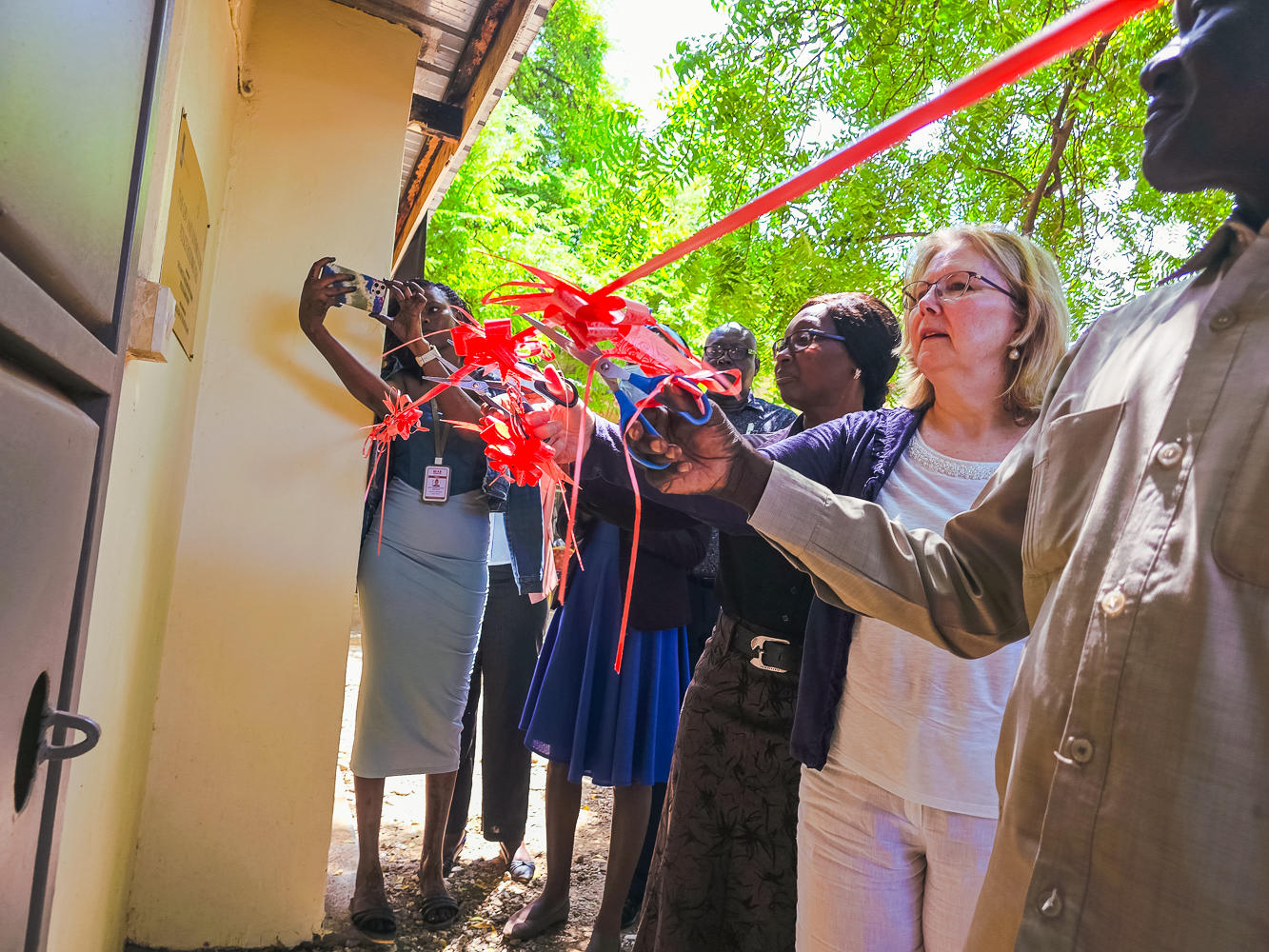 A group of people holds scissors to cut a red ribbon at an outdoor ceremony.