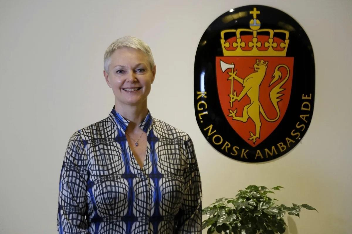 A woman with short, blonde hair smiles in front of an ambassadorial emblem on a wall.