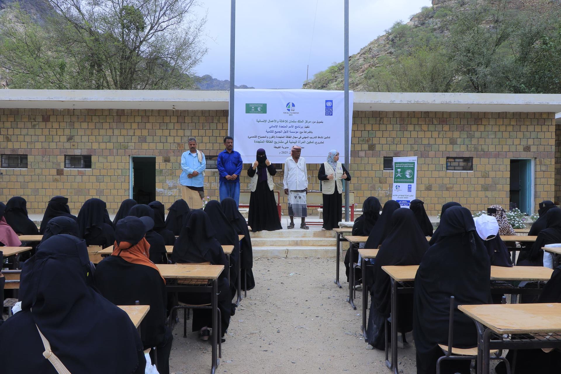 Students in black attire sitting at desks, facing speakers at an outdoor event.