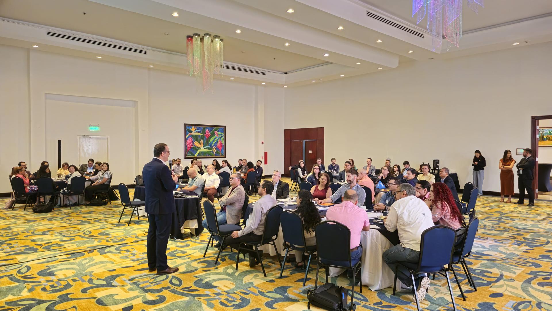 A speaker addresses a large audience seated at round tables in a brightly lit hall.