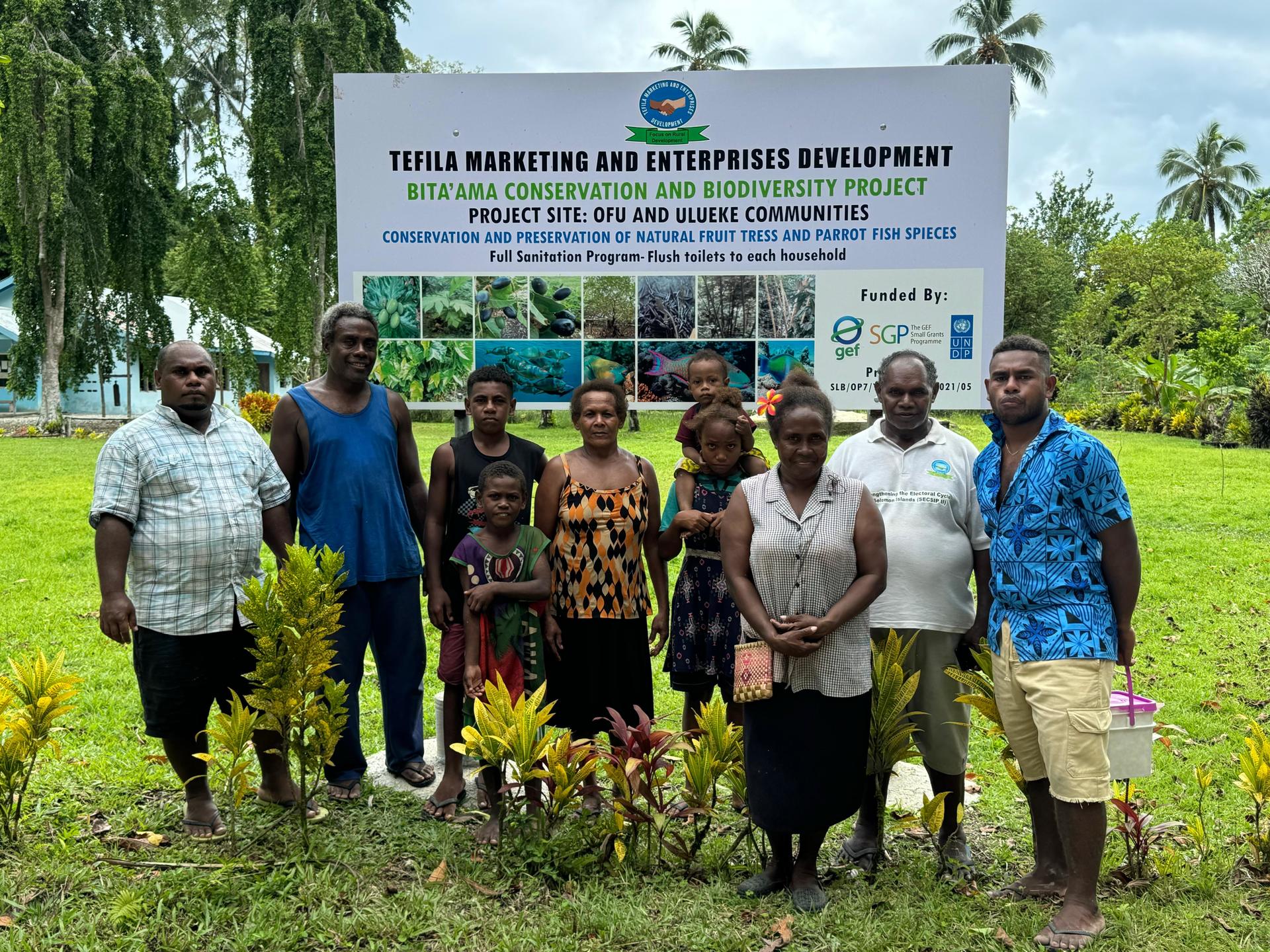 A group of diverse people stands in front of a project sign, surrounded by greenery.