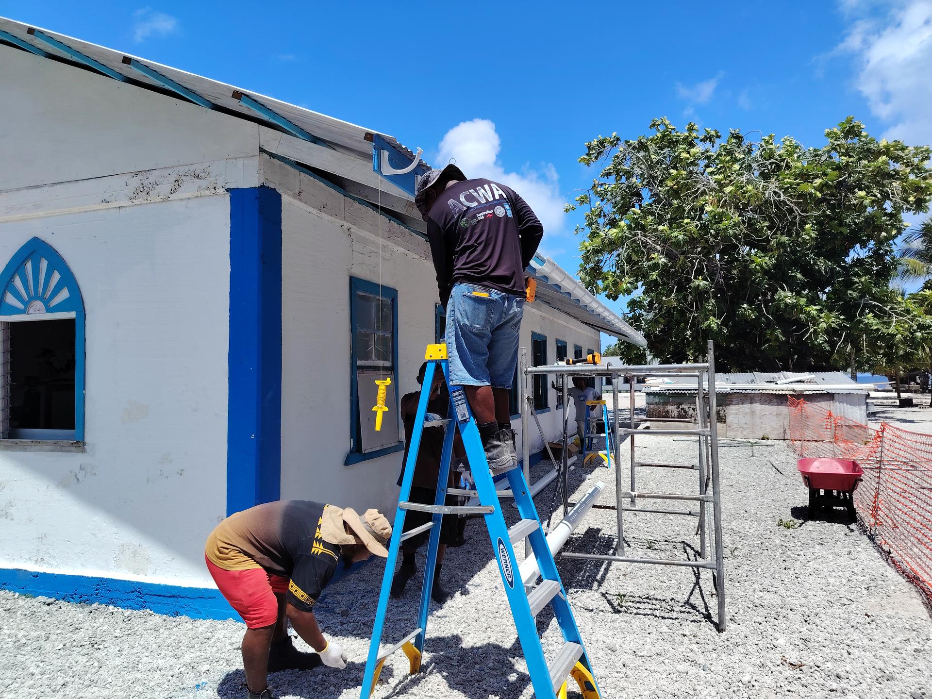 Two workers on a construction site, one on a ladder, working on a building exterior under a clear blue sky.