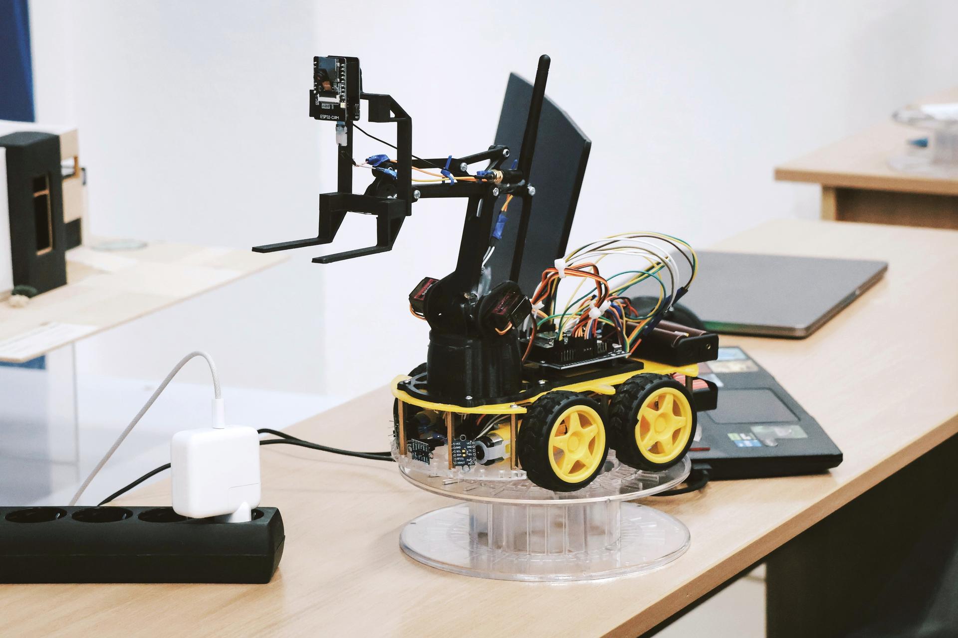 Small robot on a transparent base, equipped with wires and a monitor, on a wooden table.