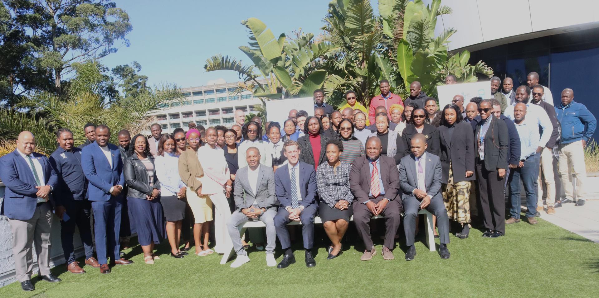 A large group of people gathered outdoors, smiling and standing on grass with greenery in the background.