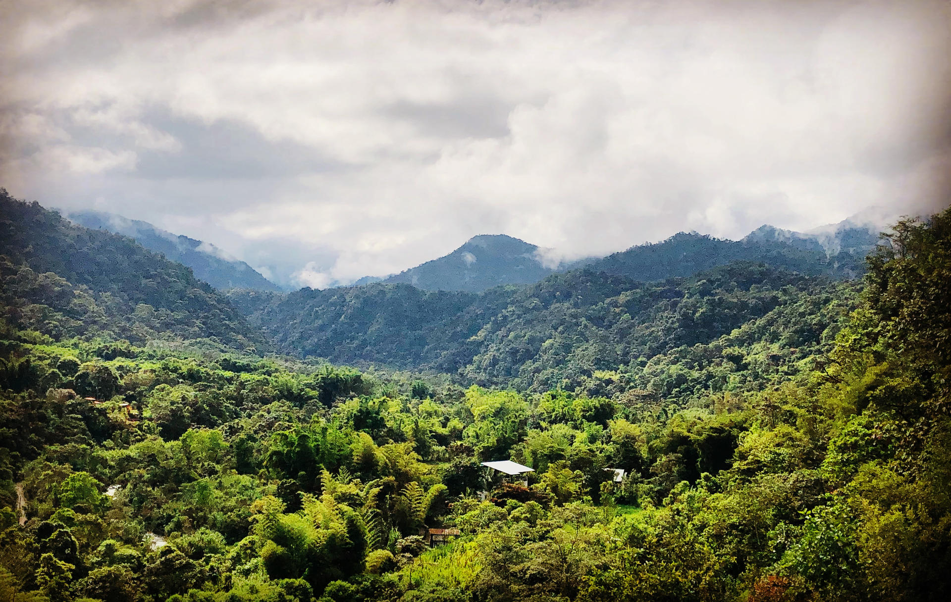 Lush green mountains under a cloudy sky, with a valley filled with trees and a small building.