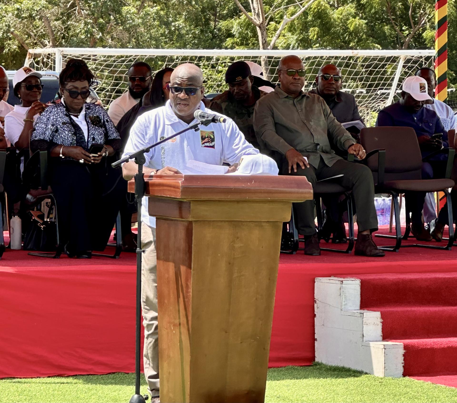 A speaker at a podium addresses an audience under a sunny sky, with people seated behind.