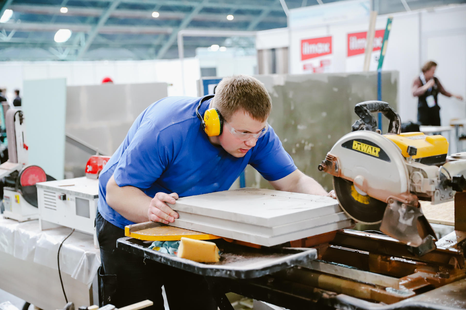 A focused worker in blue shirt and headphones measures a wooden board at a workshop.