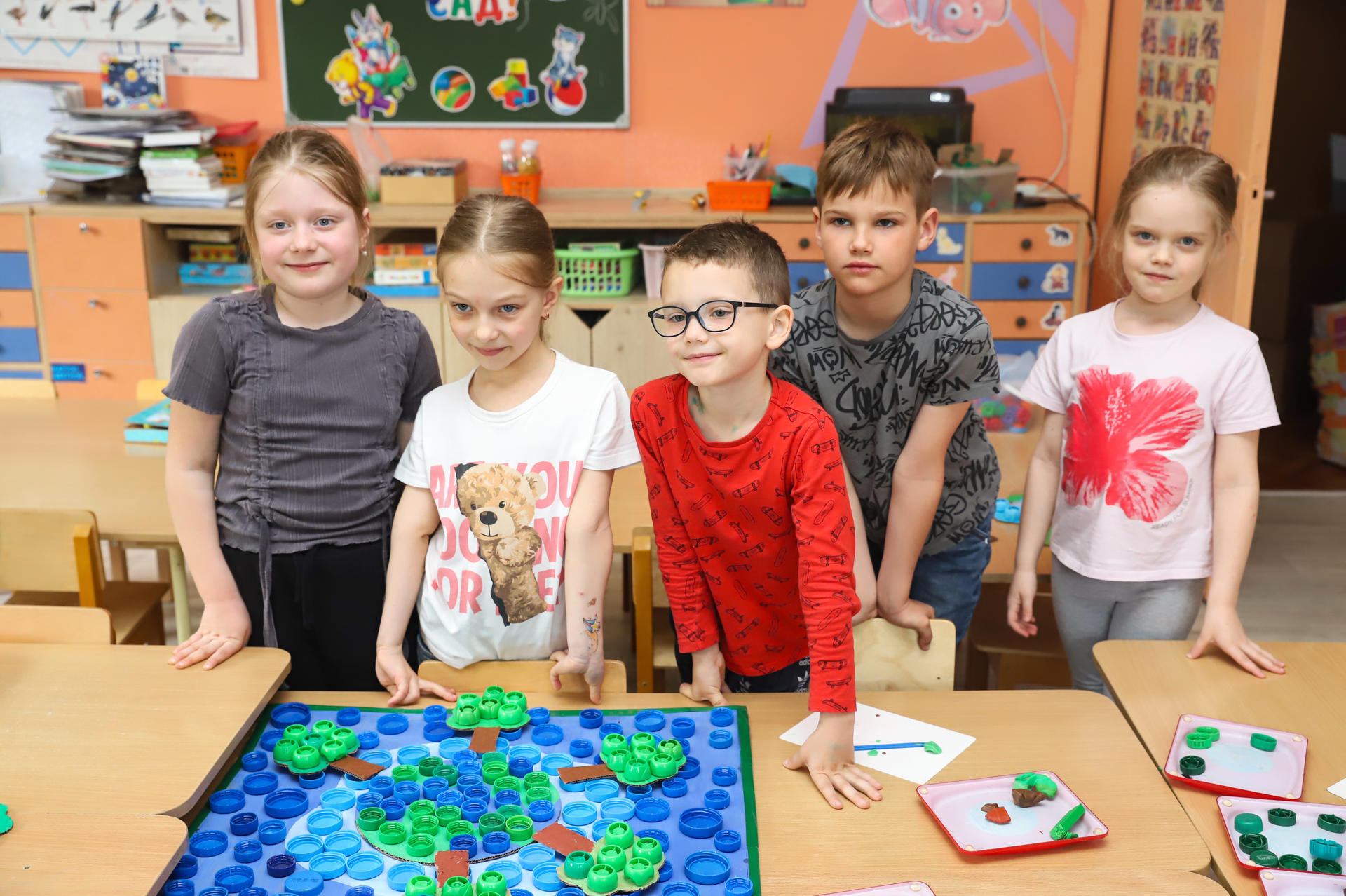 Five children stand together, smiling in a classroom with a colorful game on the table.