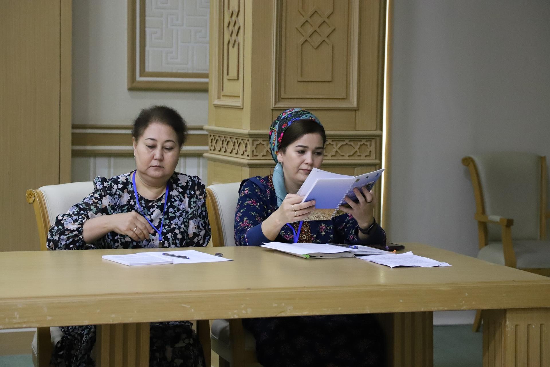 Two women sit at a table in a room, one reading a book and the other looking thoughtful.