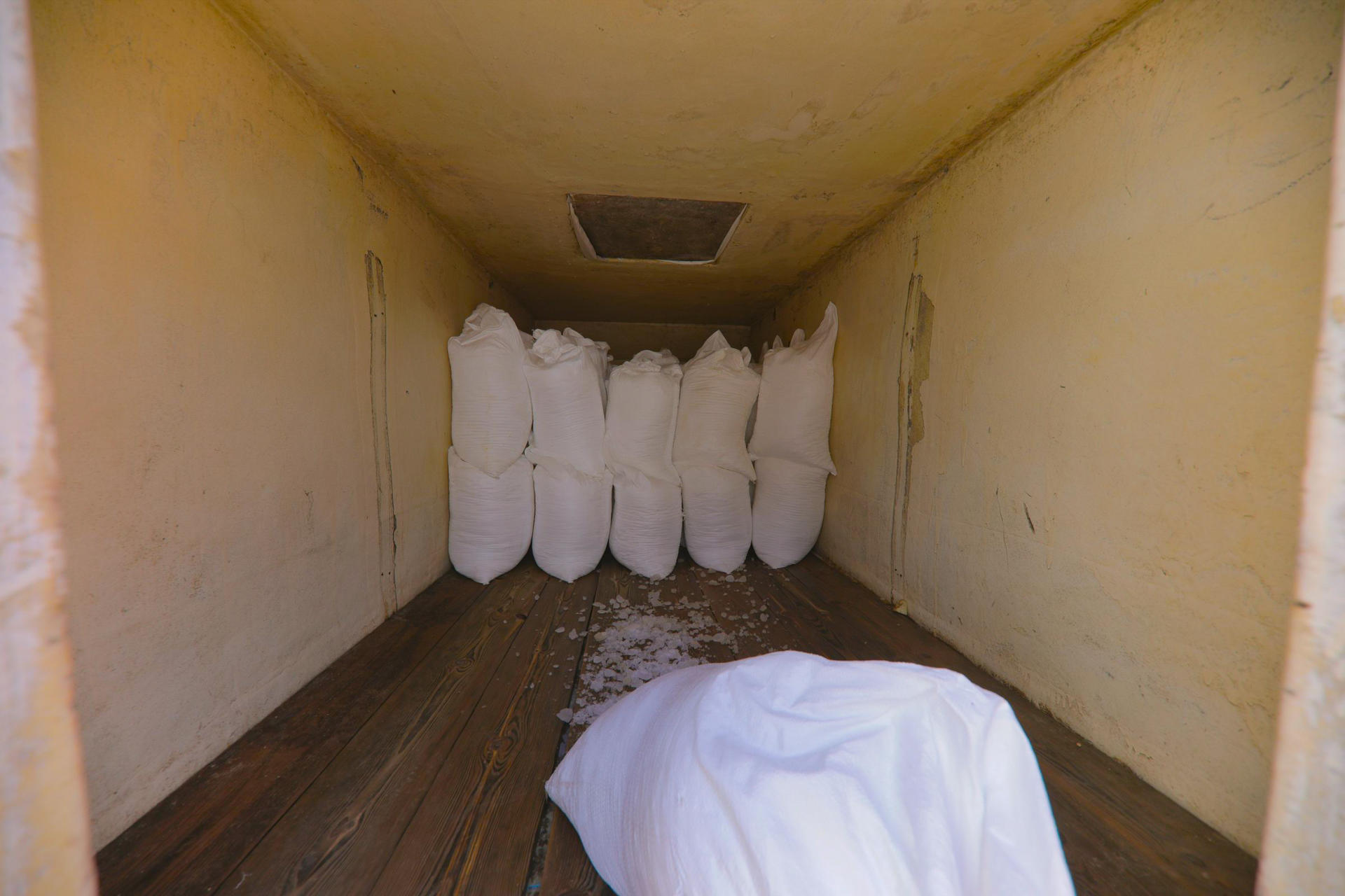 Inside a dimly lit room, several white bags are lined against the wall on a wooden floor.