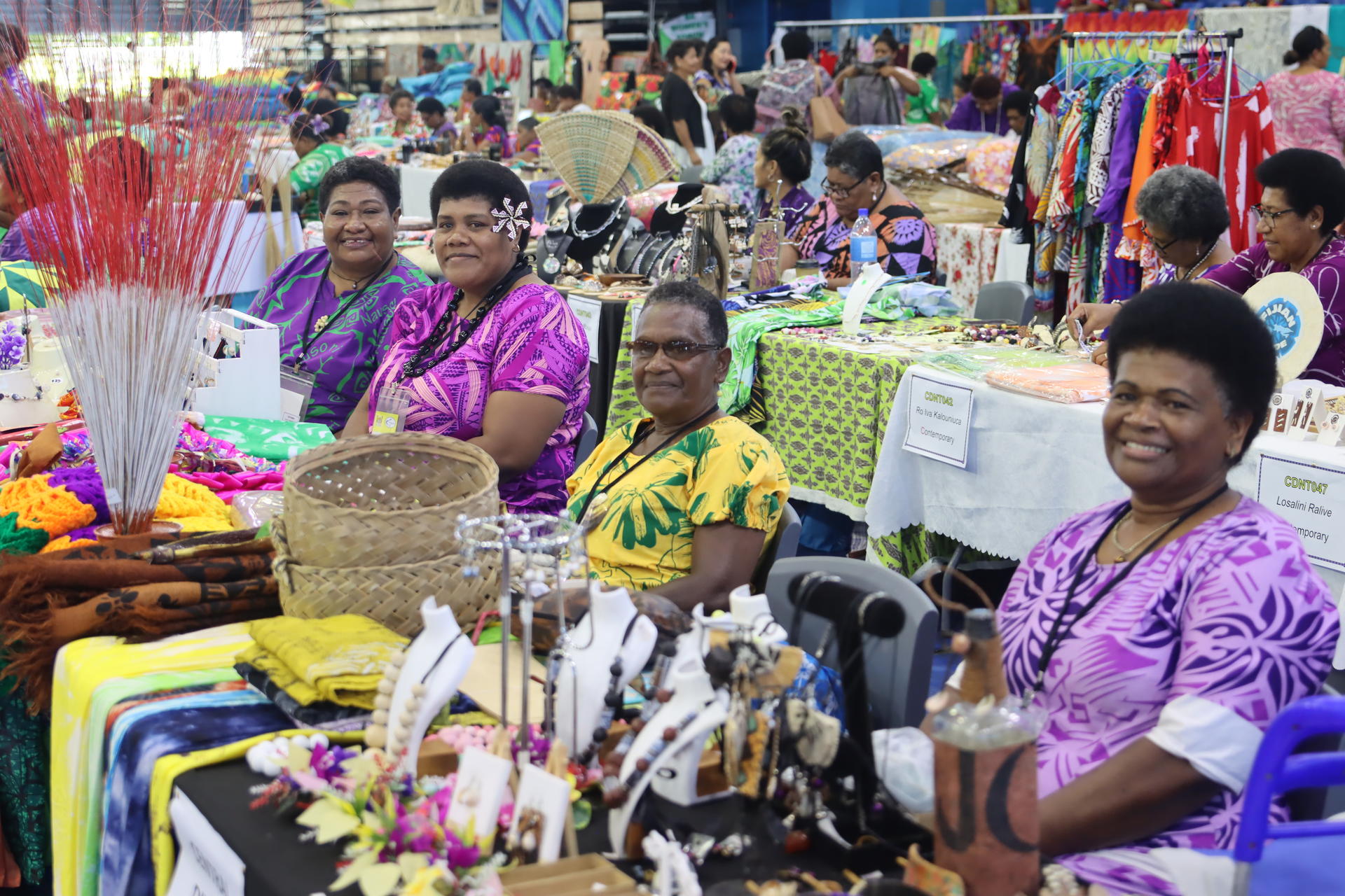 Four women sit behind tables filled with colorful crafts and decorations at a market.