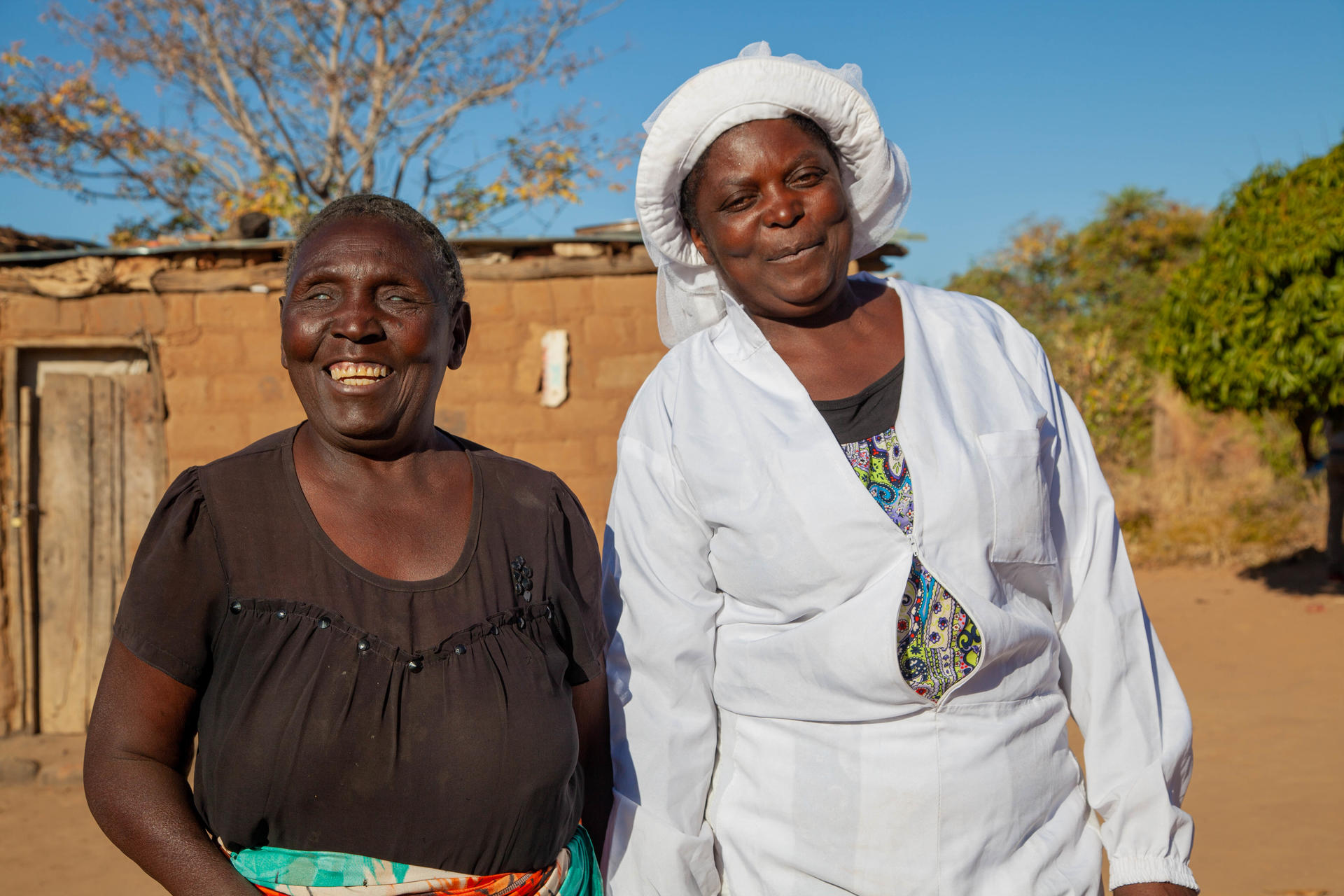 Two smiling women stand outdoors, wearing traditional attire, with a rustic background.