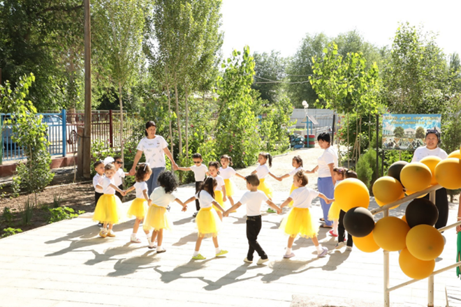 Children in yellow skirts dance in a circle outdoors, surrounded by greenery and balloons.