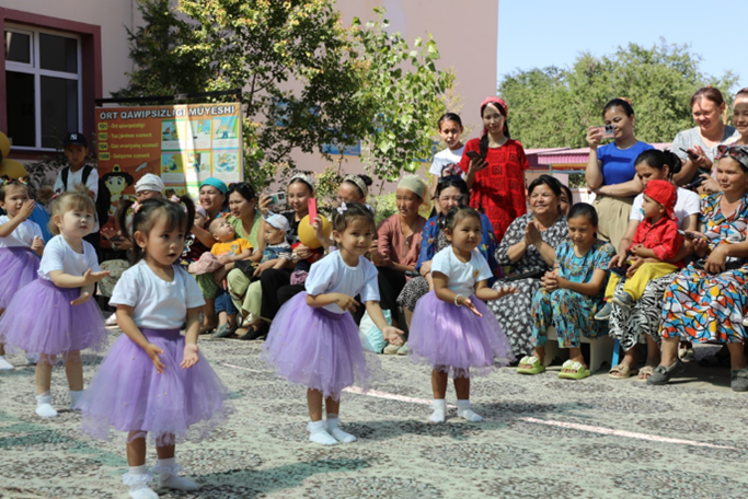 Children in purple tutus perform in front of a colorful audience outdoors.