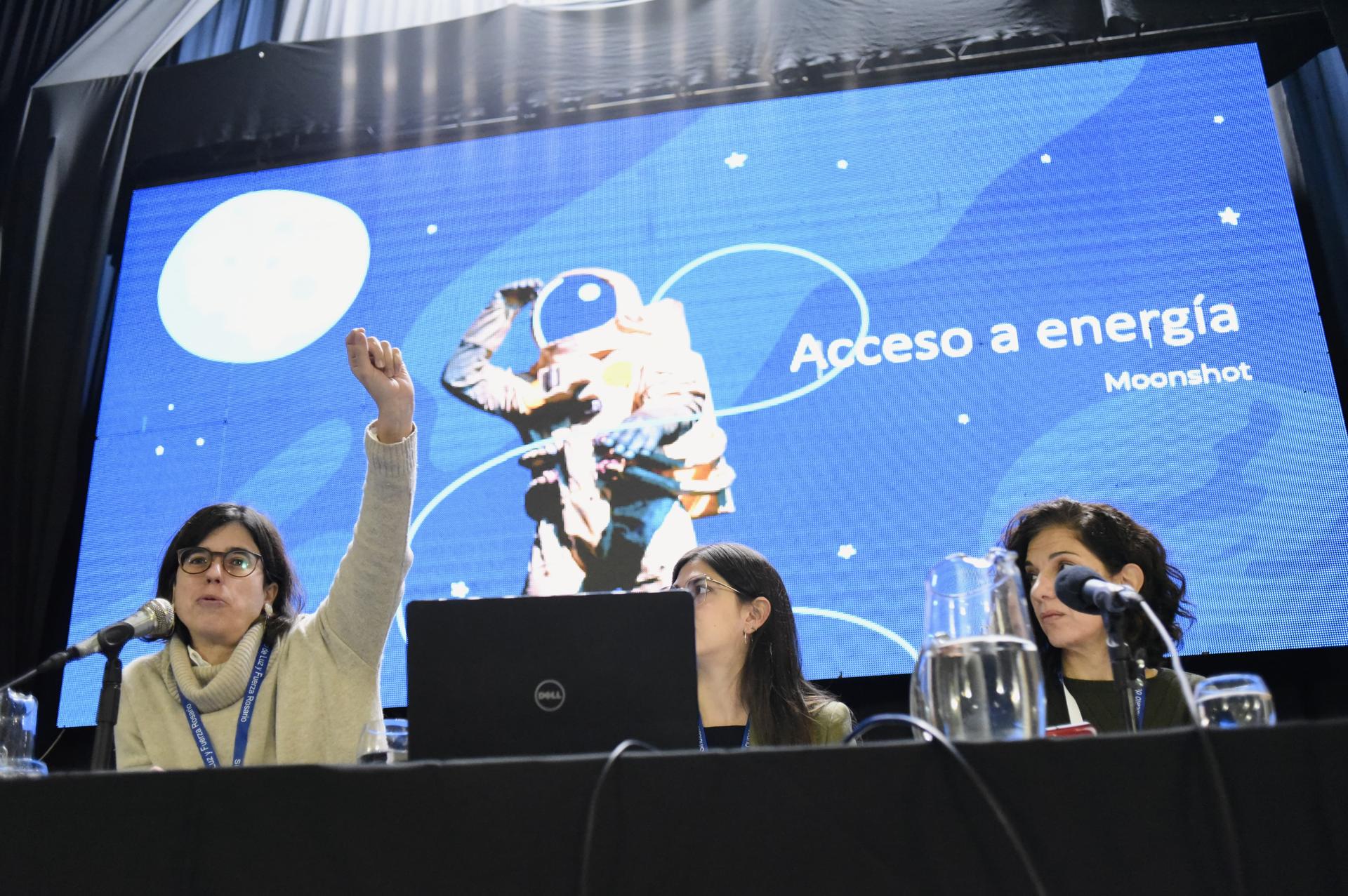 Tres mujeres en una mesa de conferencia, una con una mano levantada, con una presentación con gráficos del espacio en el fondo.
