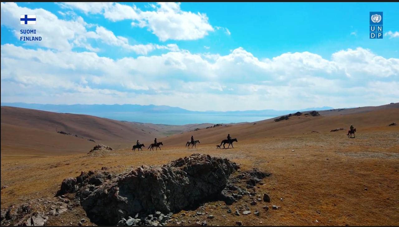 Several horseback riders traverse a vast, grassy landscape under a blue sky with clouds.