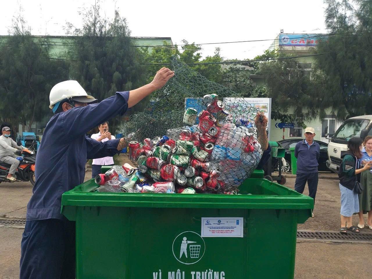 A person wearing a helmet deposits crumpled cans into a green recycling bin.